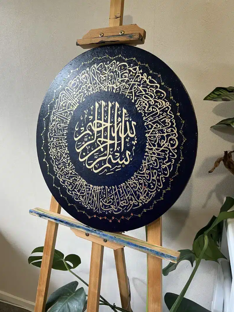 The Verse of The Throne, a round artwork with intricate gold Arabic calligraphy on a dark blue background, is displayed on a wooden easel with green plants nearby.