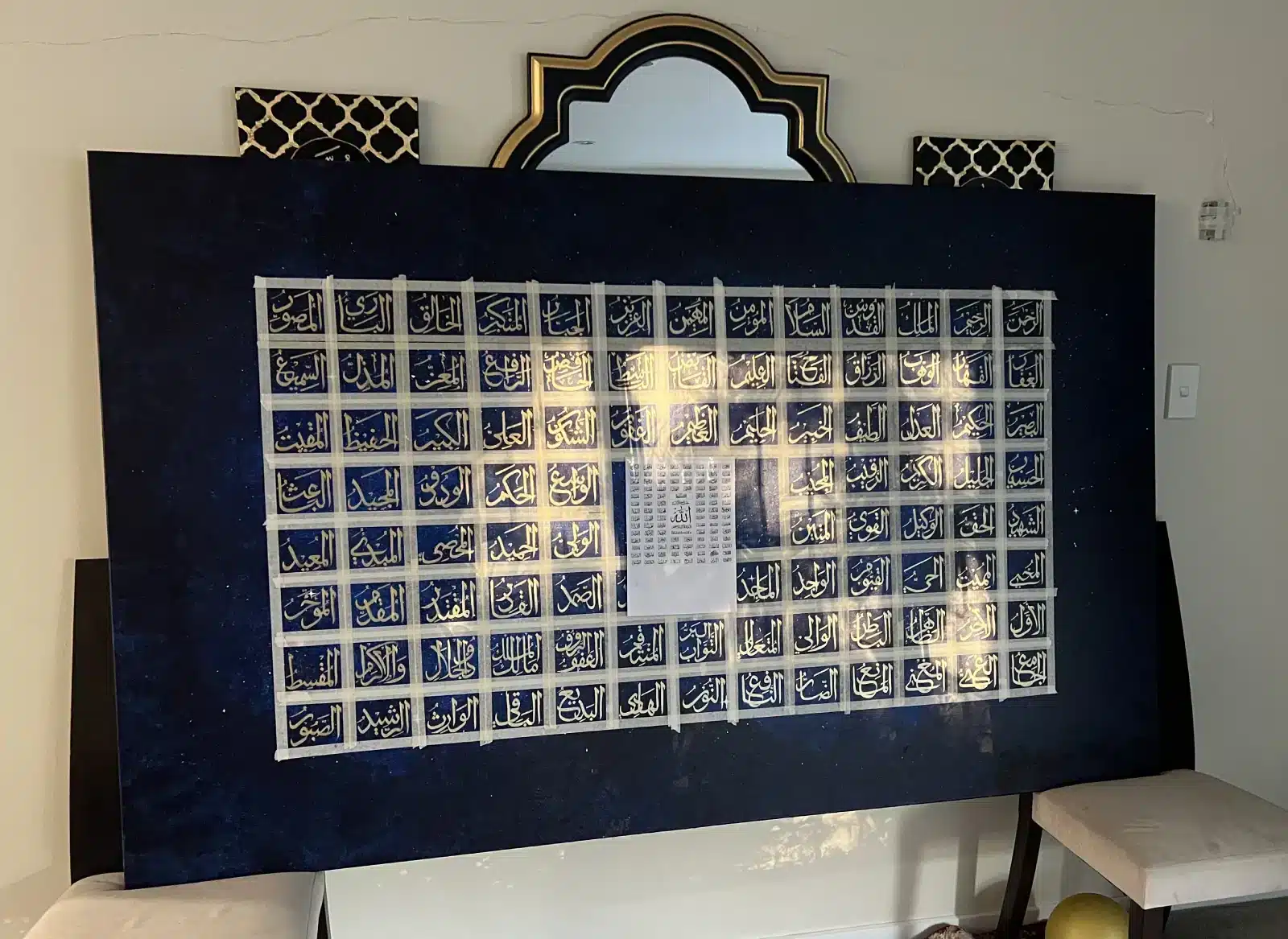 The Asma al-Husna Artwork, a large rectangular piece featuring white Arabic calligraphy on a blue background, rests against a wall and partially covers a mirror. A paper chart is taped at its center, with two stools placed nearby.