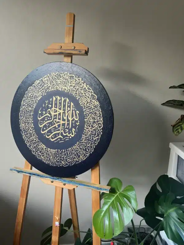 The Verse of The Throne, a round black canvas featuring intricate gold Arabic calligraphy, is showcased on a wooden easel with green houseplants nearby, set against a plain light-colored wall.